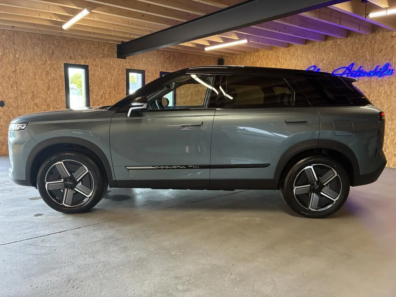 SUV JAECOO J7 2026 gris olive avec toit noir, vu de profil côté gauche dans un intérieur en bois.