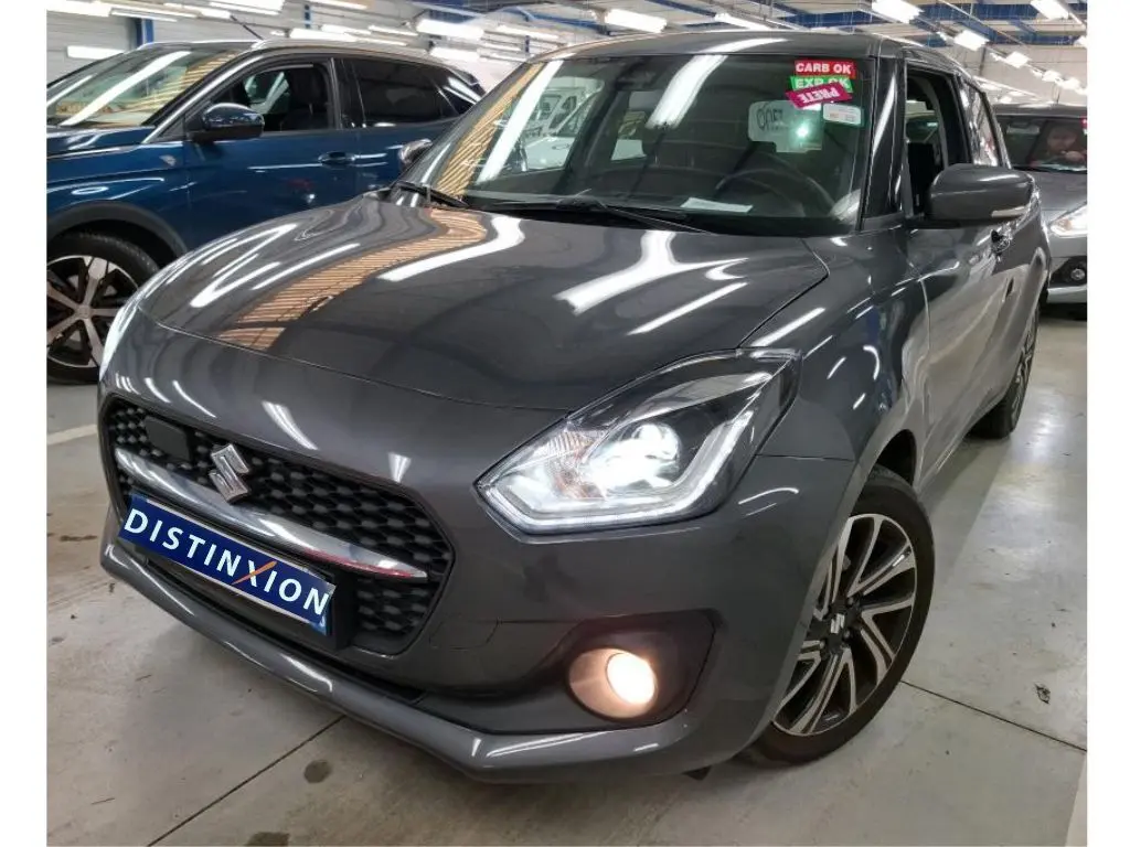 Suzuki Swift grise vue 3/4 avant droit, phares et antibrouillard allumés en intérieur showroom.
