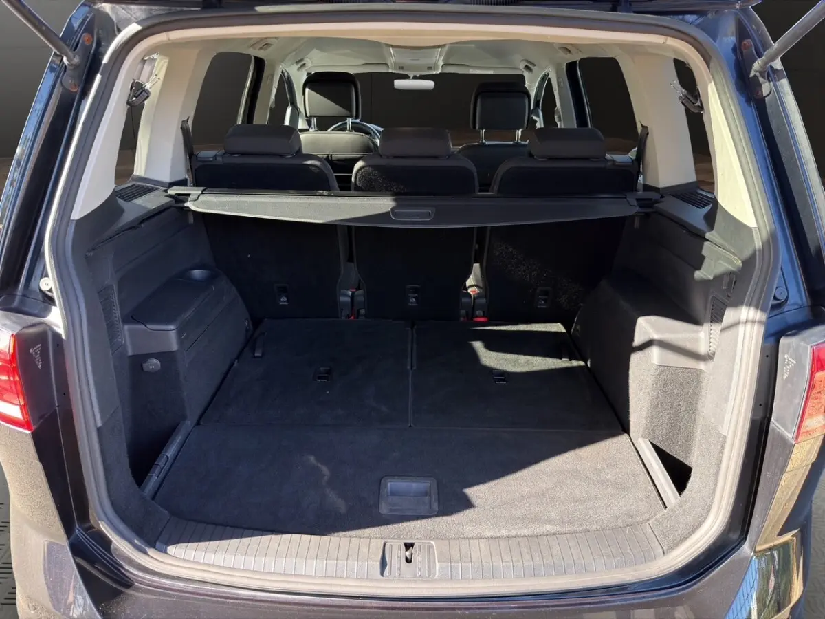 Coffre ouvert vu de l'arrière d'un Volkswagen Touran noir, montrant les sièges rabattus et la tablette de coffre.