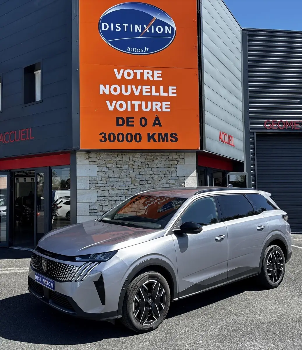 Peugeot 5008 gris clair en 3/4 avant droit, avec calandre distinctive et jantes noires, devant un bâtiment commercial.