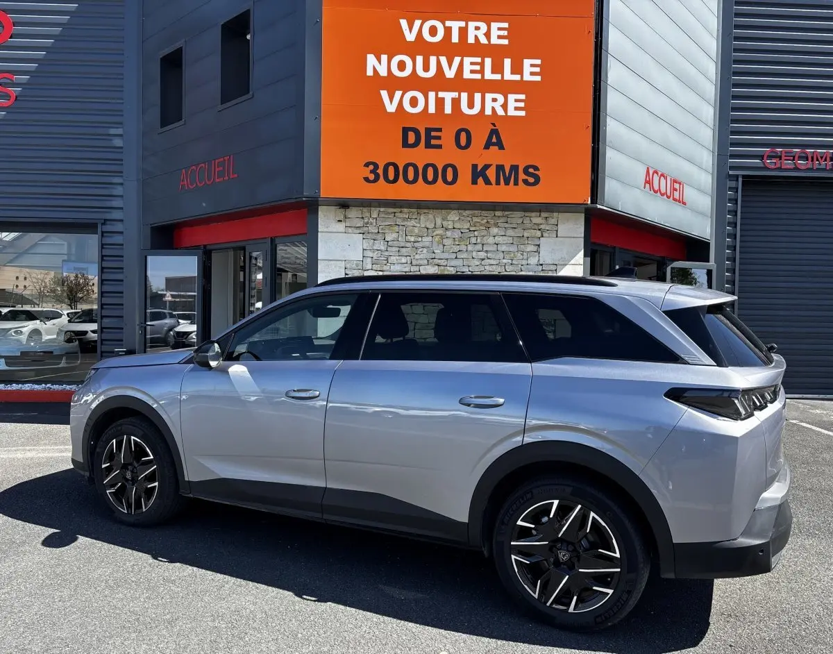 Vue de profil côté gauche d’un Peugeot 5008 gris clair 2025 garé devant un bâtiment avec panneau orange.