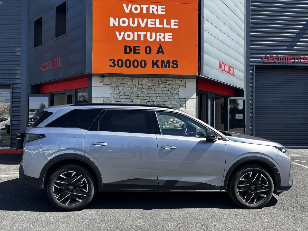 Peugeot 5008 gris clair vue de profil côté gauche, SUV hybride 2025 garé devant un bâtiment avec panneau orange.