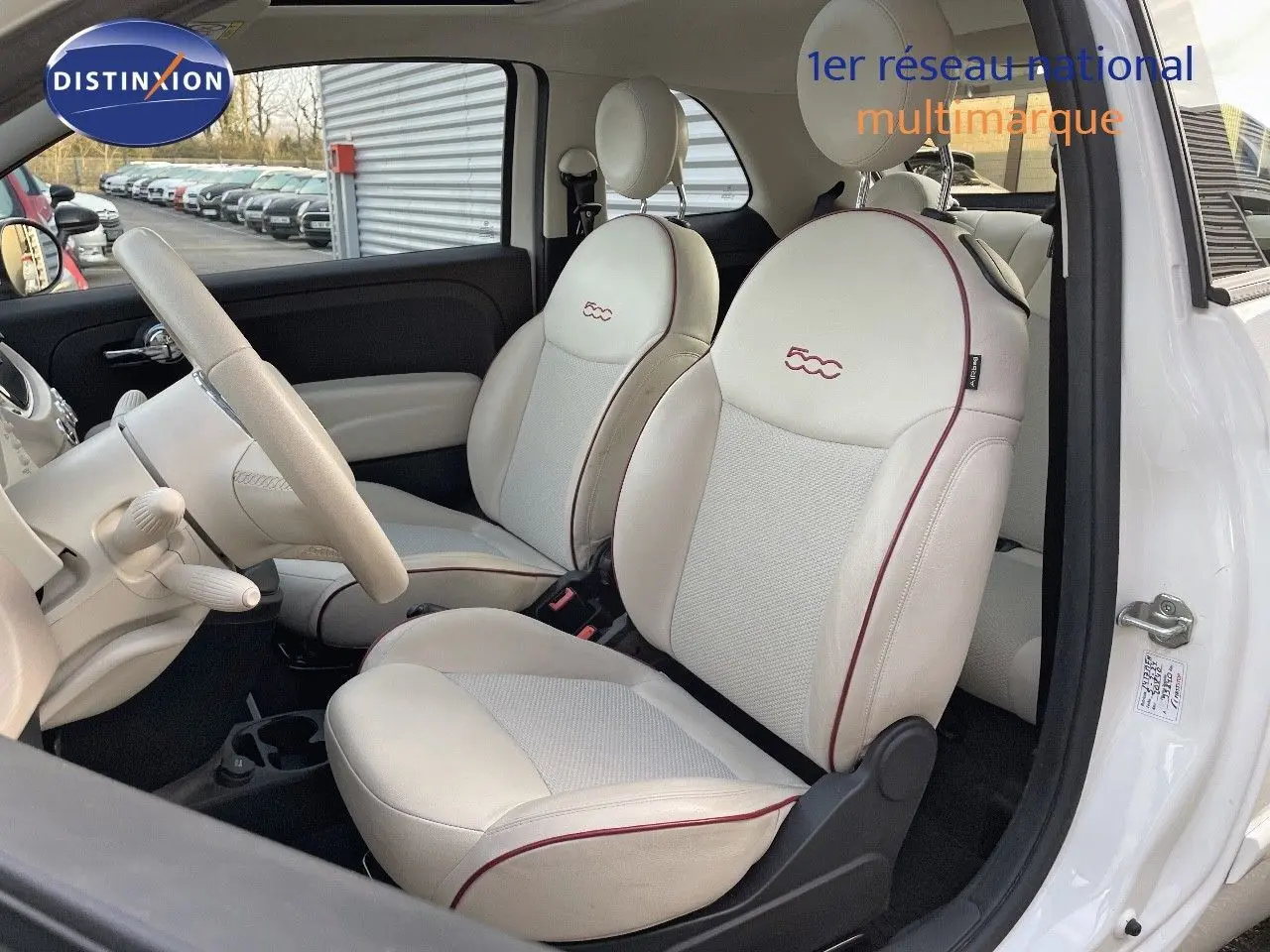 Vue intérieure côté conducteur de la Fiat 500 blanche 1.0 Hybrid 70ch DolceVita avec sièges cuir blanc et surpiqûres rouges.