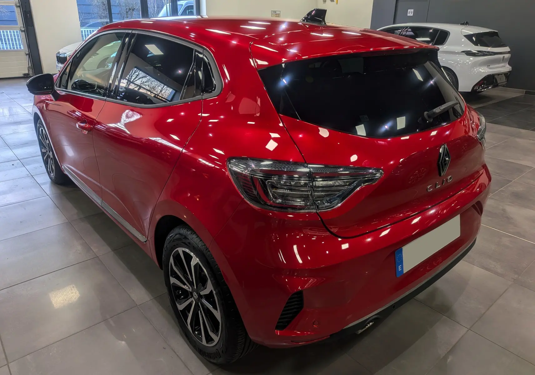 Vue 3/4 arrière droite d'une Renault Clio V rouge flamme avec vitres teintées et jantes alliage bicolores en showroom.
