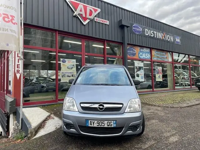 Vue de face d'une Opel Meriva grise de 2010 stationnée devant un garage avec logo AD Expert visible.