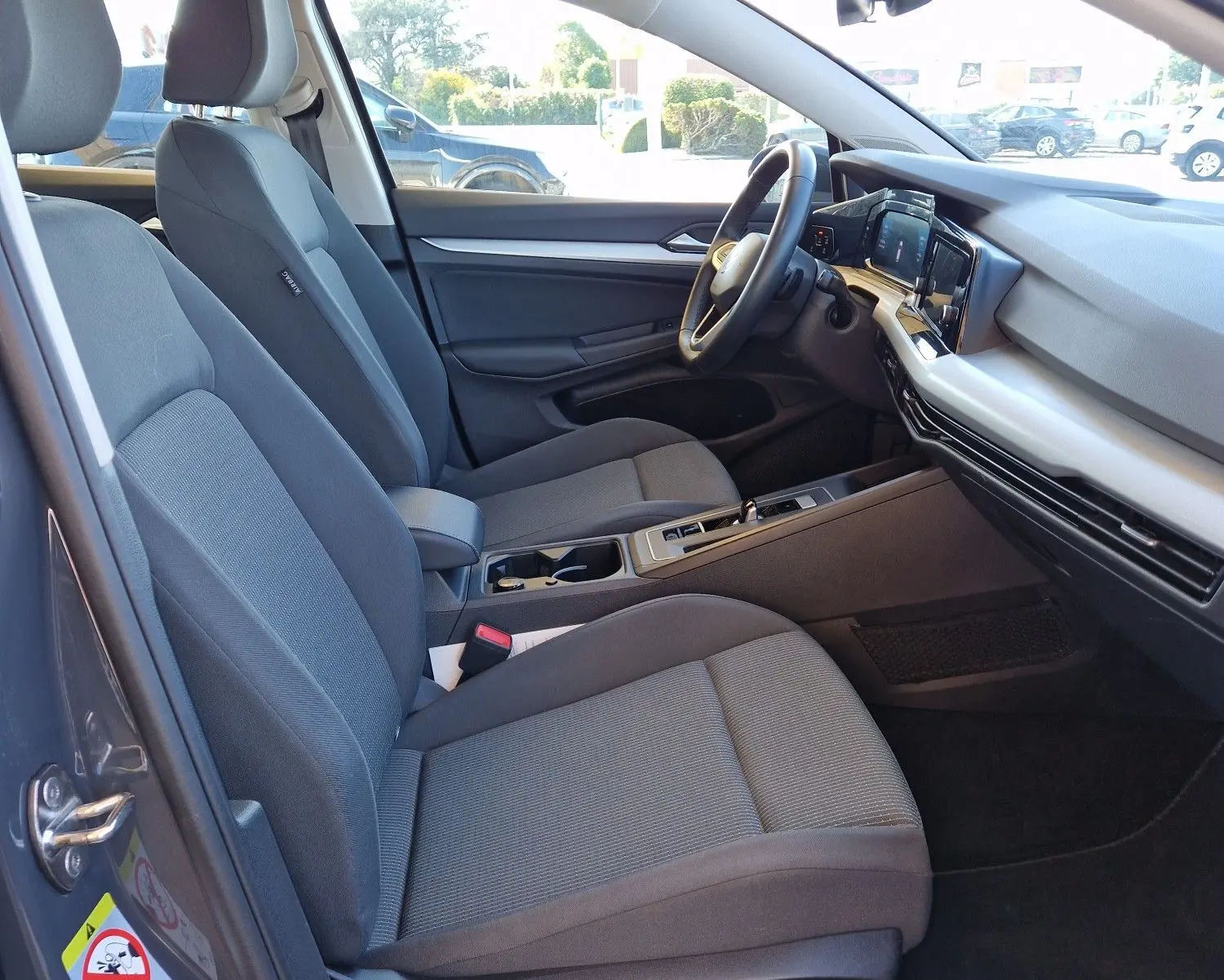 Vue intérieure côté conducteur de la Volkswagen Golf A8 2023 gris Urano, montrant sièges tissu et tableau de bord moderne.