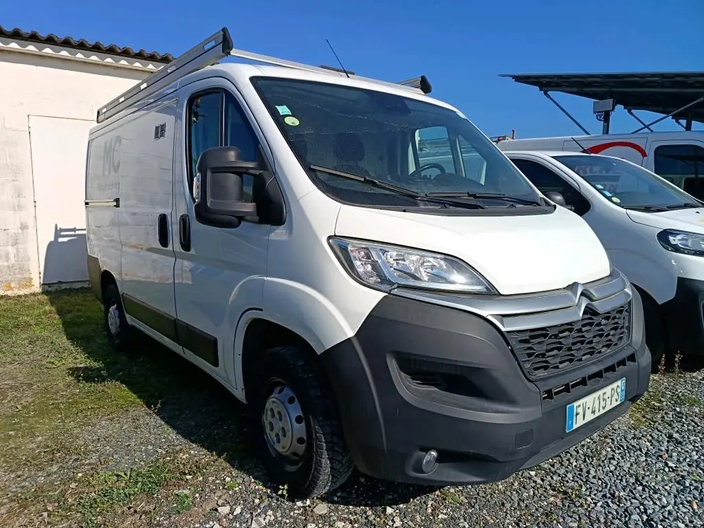 Vue 3/4 avant d'un utilitaire blanc Citroën Jumper L1H1 avec galerie de toit et pare-chocs noir sur un sol gravillonné.