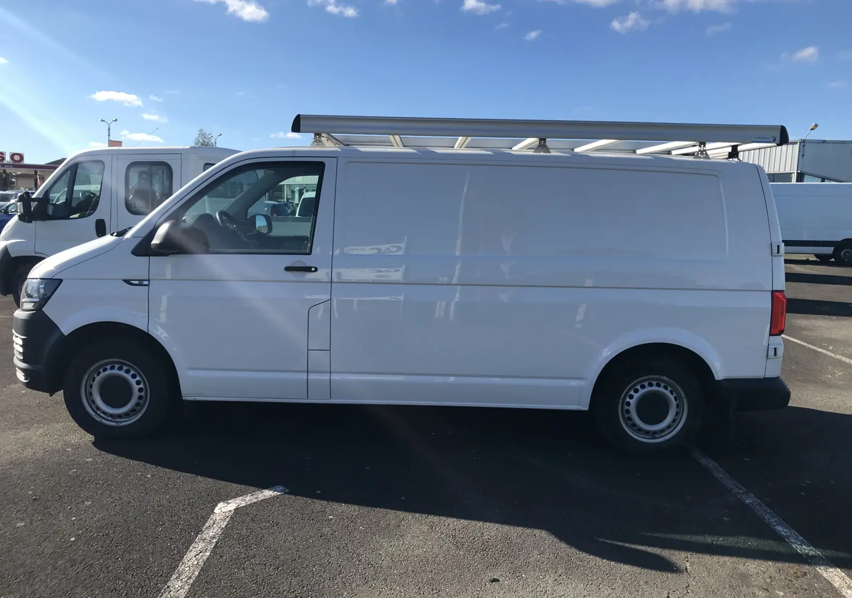 Vue latérale côté gauche d'un Volkswagen Transporter blanc avec galerie de toit sur un parking ensoleillé.