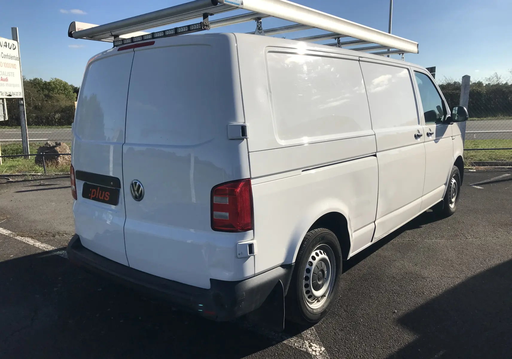 Vue 3/4 arrière droite d'un Volkswagen Transporter blanc équipé d'un porte-échelle sur le toit, stationné en extérieur.