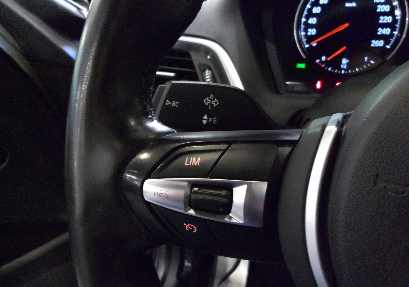 Gros plan sur les commandes au volant noir de la BMW Série 1 F21 LCI 118i, avec tableau de bord éclairé en arrière-plan.