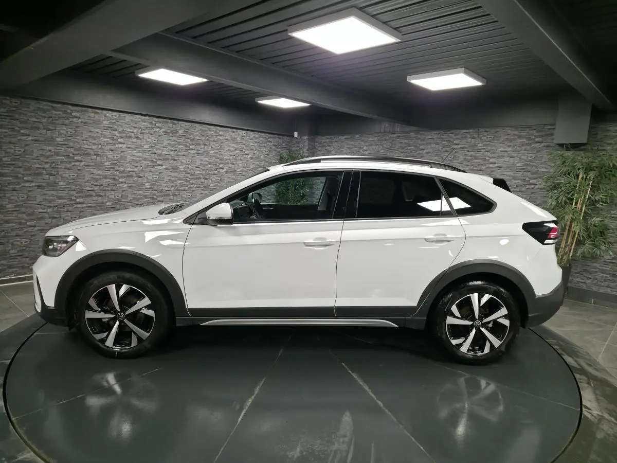 Profil droit du Volkswagen Taigo blanc 2024 avec jantes alliage 17 pouces et barres de toit noires dans un showroom.