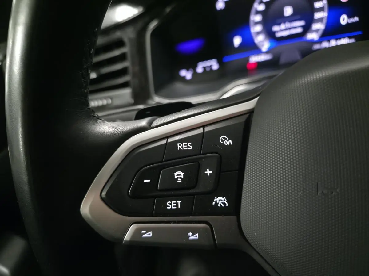 Gros plan sur les commandes du régulateur de vitesse au volant cuir noir du Volkswagen Taigo blanc, tableau de bord numérique en arrière-plan.