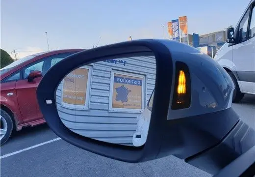 Rétroviseur droit d'une Volkswagen ID.3 blanc glacier avec clignotant allumé, vue sur un parking extérieur.