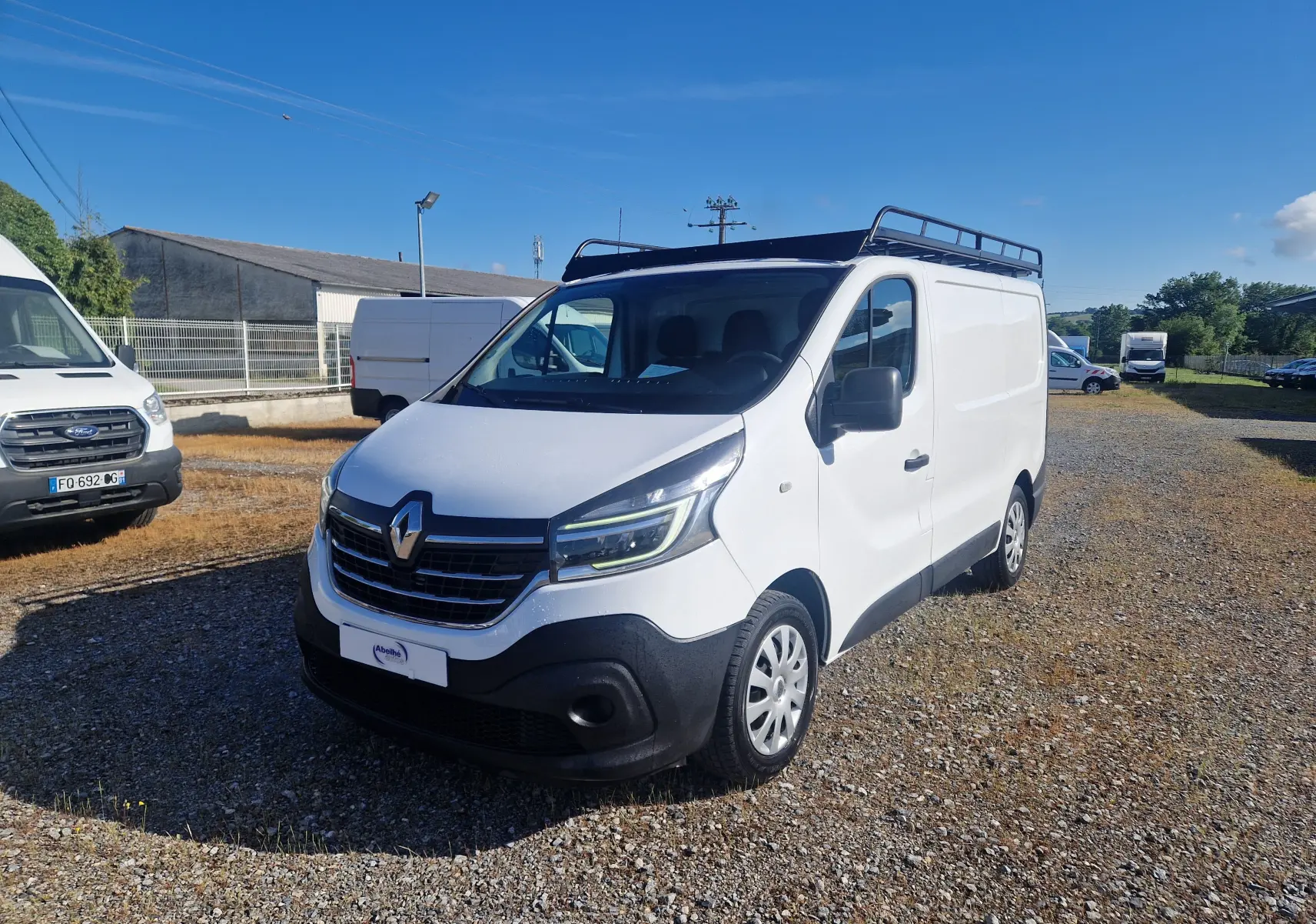 Vue 3/4 avant droite du Renault Trafic Fourgon blanc avec galerie de toit sur un terrain extérieur ensoleillé.