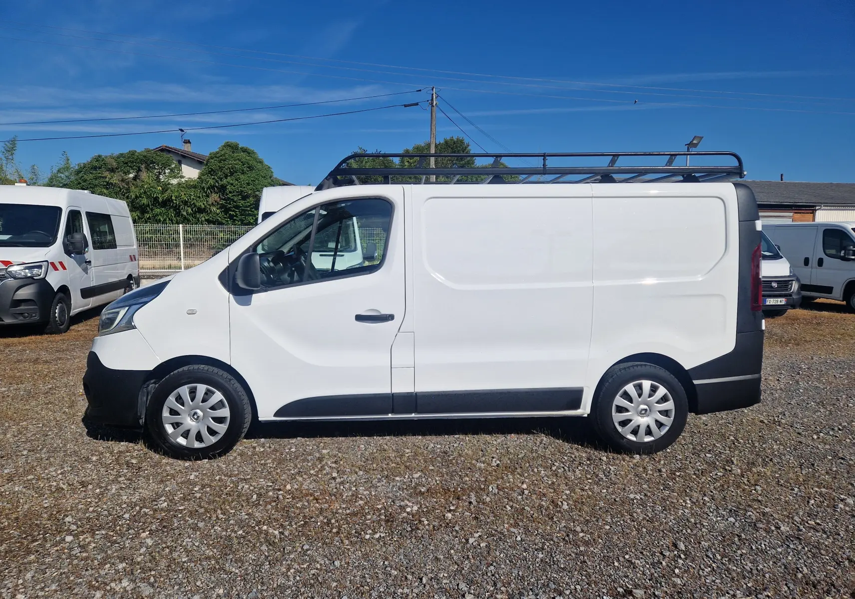 Profil droit d’un fourgon blanc Renault Trafic L1H1 2021 avec galerie de toit noire, stationné sur un sol gravillonné.