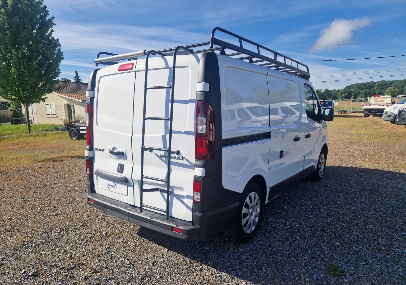 Vue 3/4 arrière droite d’un Renault Trafic Fourgon blanc avec échelle et galerie de toit noire sur un terrain extérieur.