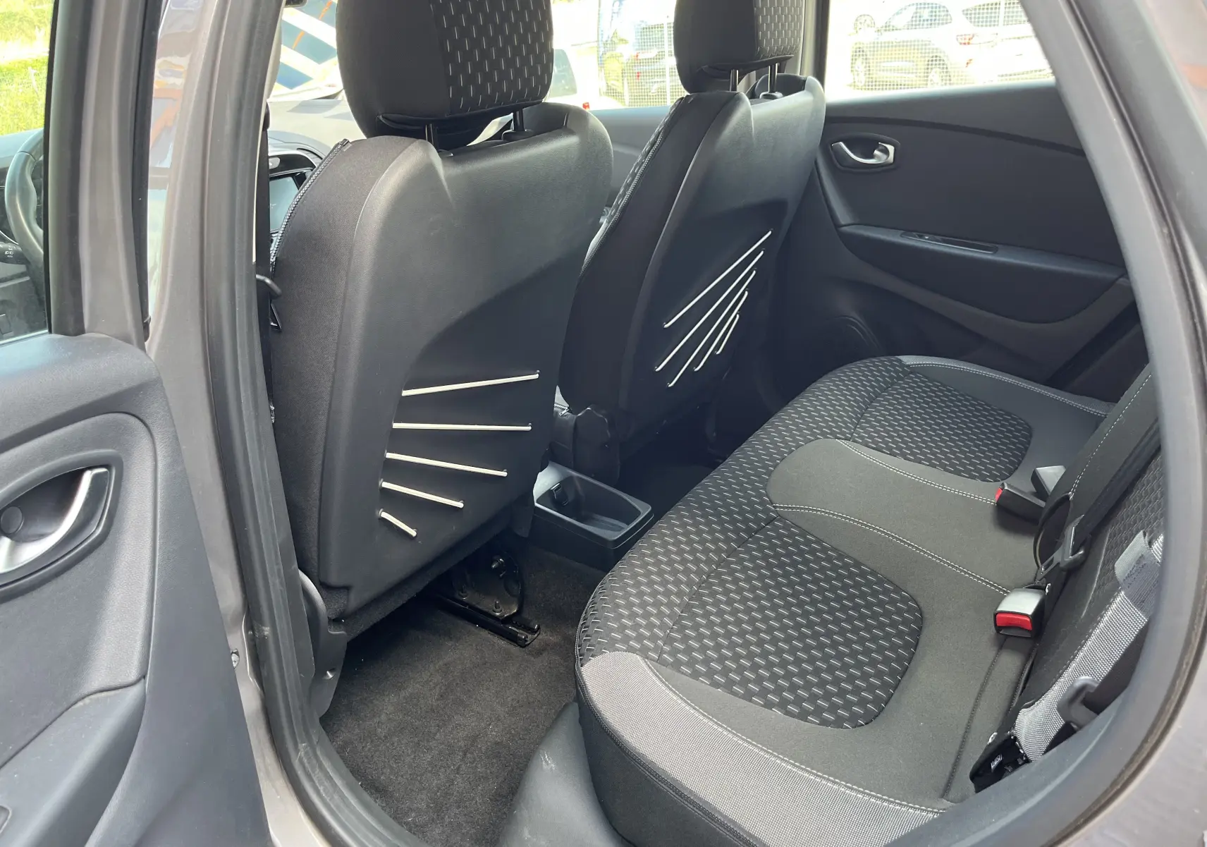 Vue intérieure arrière côté gauche du Renault Captur gris 2019, montrant les sièges arrière en tissu noir à motifs et les dossiers avant avec inserts décoratifs.