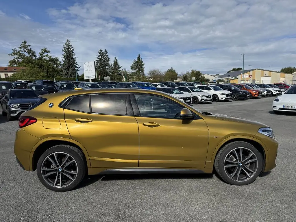 Profil côté gauche d'un BMW X2 jaune métallisé avec jantes M sport noires sur un parking extérieur.