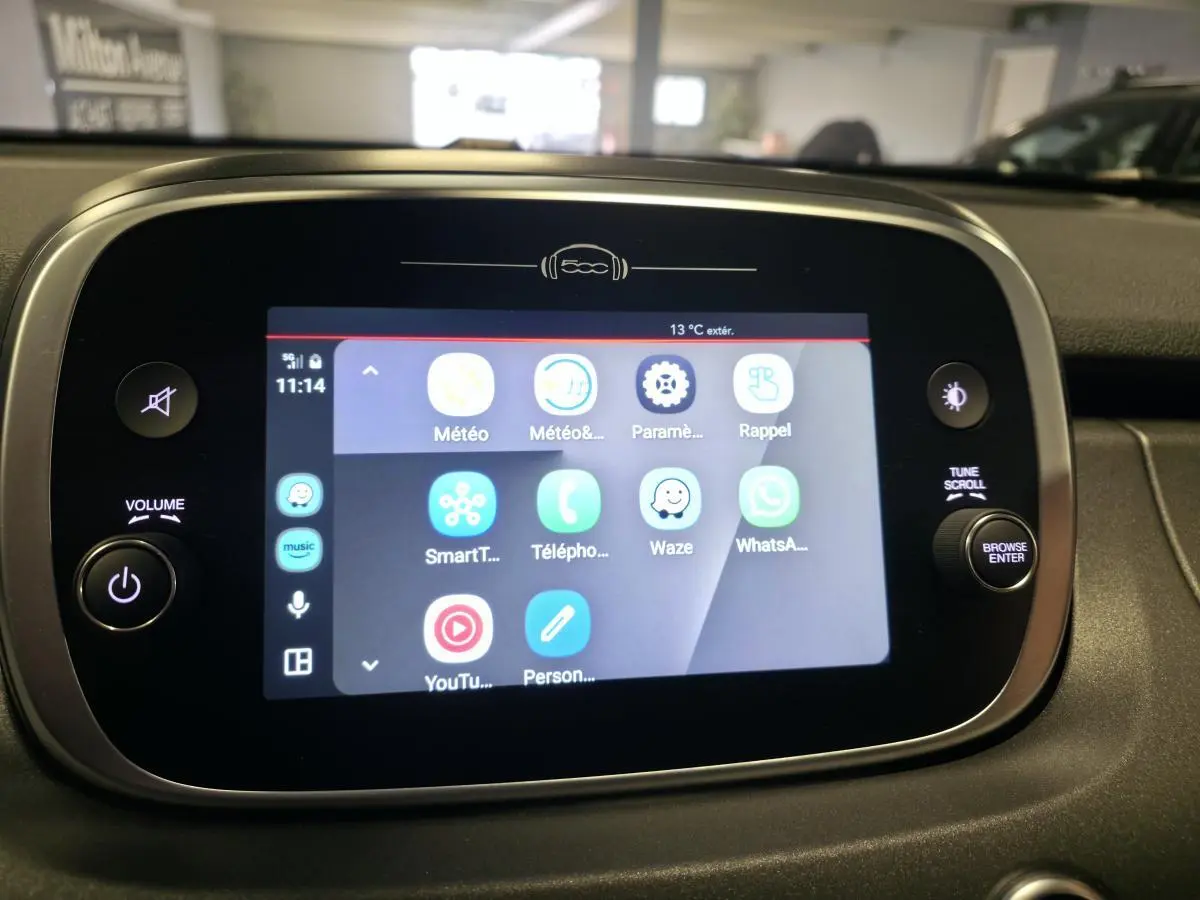 Écran tactile multimédia de la Fiat 500X 2024 affichant applications connectées, entouré de commandes volume et navigation.
