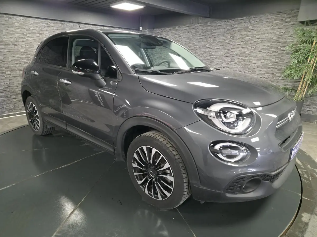 Fiat 500X gris vue 3/4 avant droit, jantes alliage 17'', phares LED et rétroviseurs noirs dans un intérieur moderne.