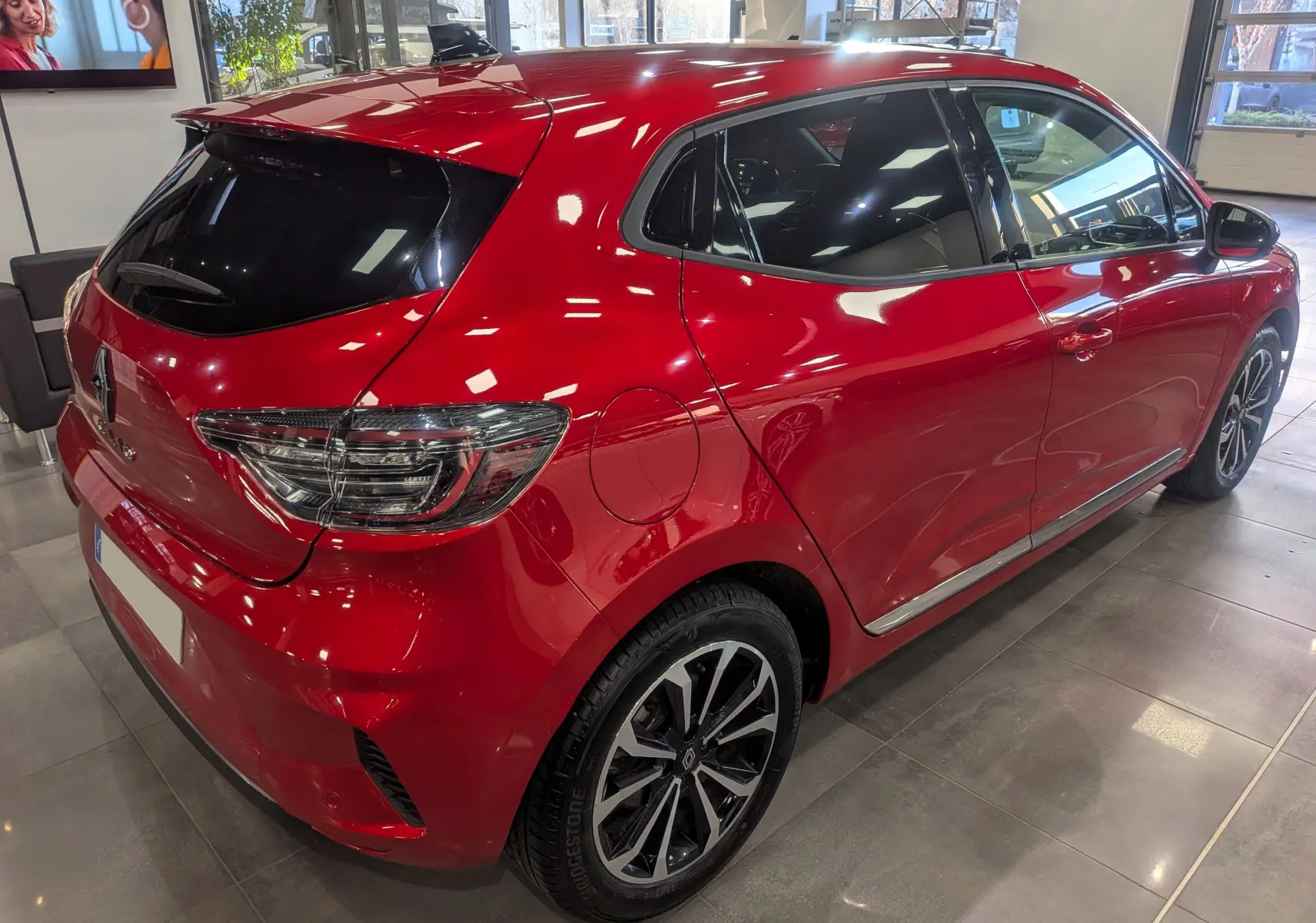 Vue 3/4 arrière droite d'une Renault Clio V Rouge Flamme avec jantes alliage et vitres teintées en intérieur showroom.