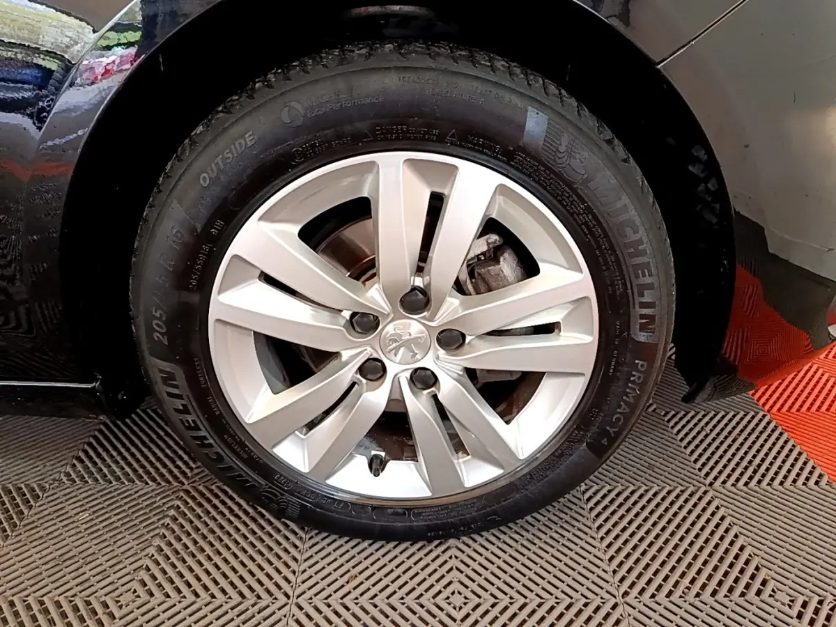 Gros plan sur la roue avant droite d’une Peugeot 308 noire, jante alliage argentée avec logo lion central.