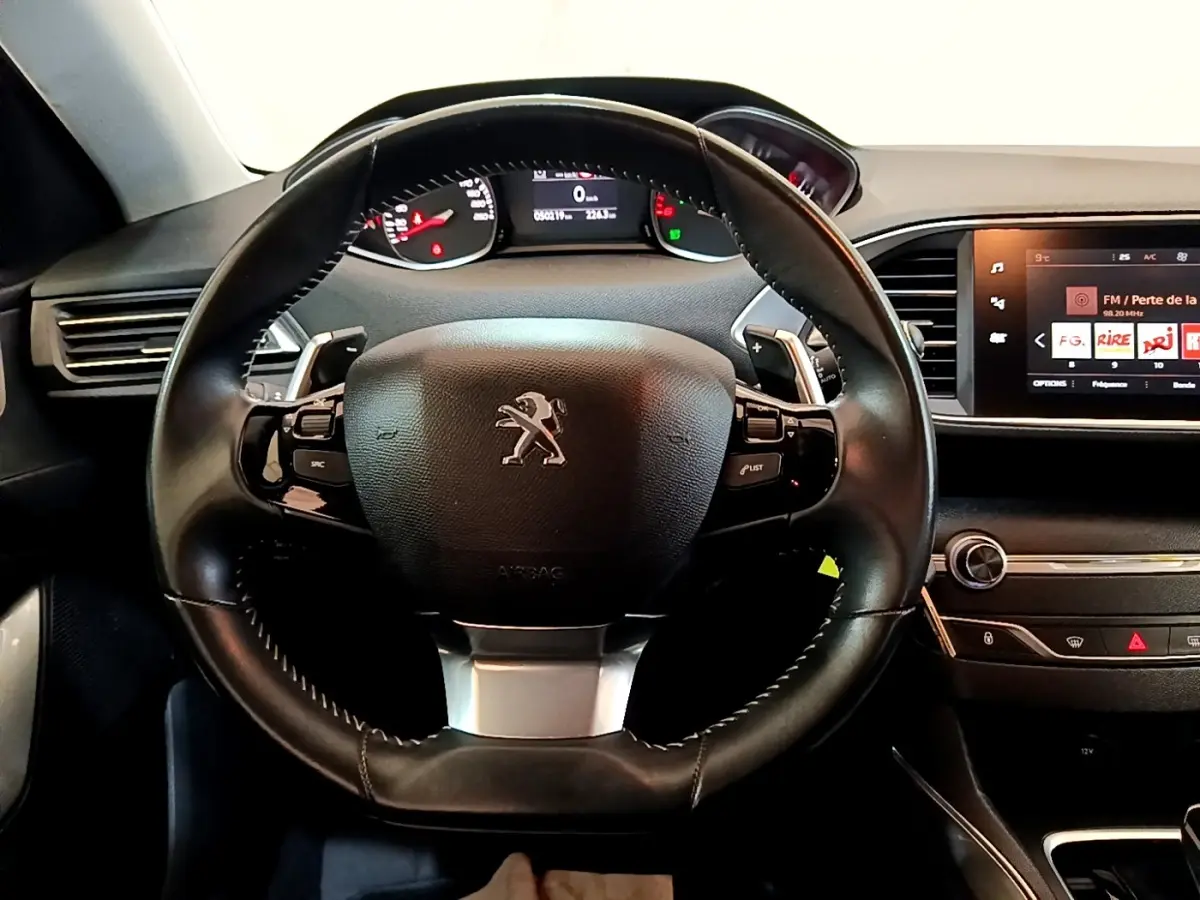 Vue intérieure centrée sur le volant cuir noir de la Peugeot 308, avec tableau de bord et écran tactile visible à droite.