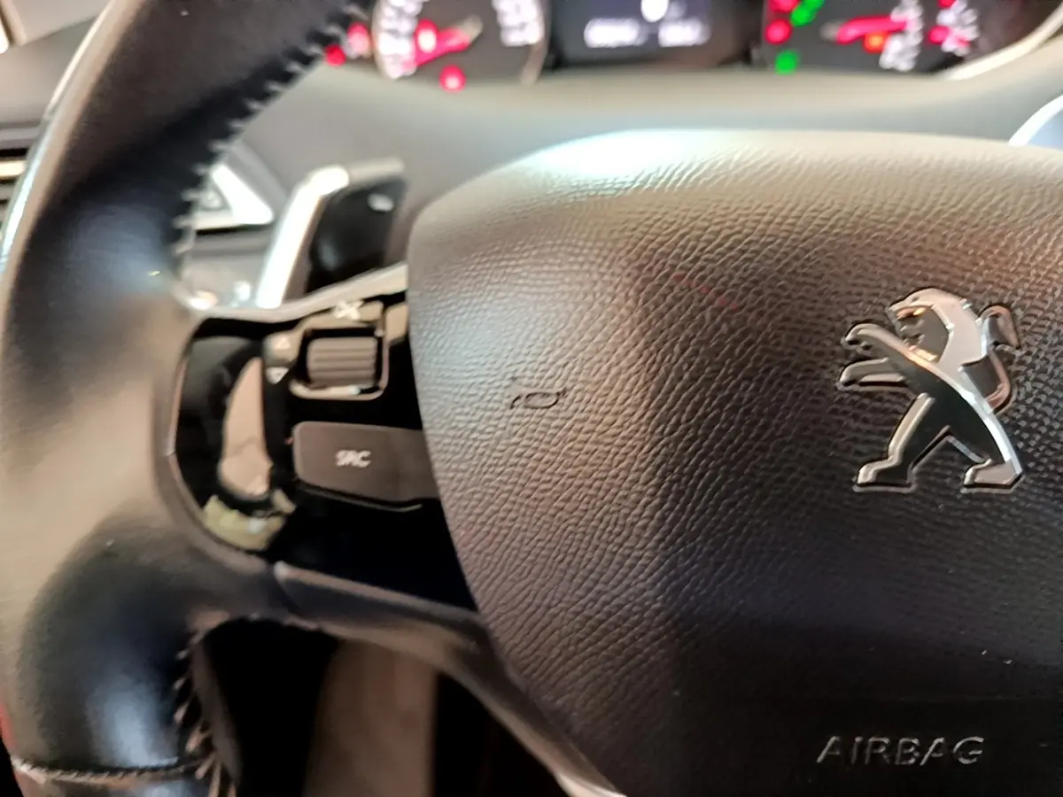 Gros plan sur le volant cuir noir de la Peugeot 308 avec logo lion et commandes intégrées, tableau de bord flou en arrière-plan.