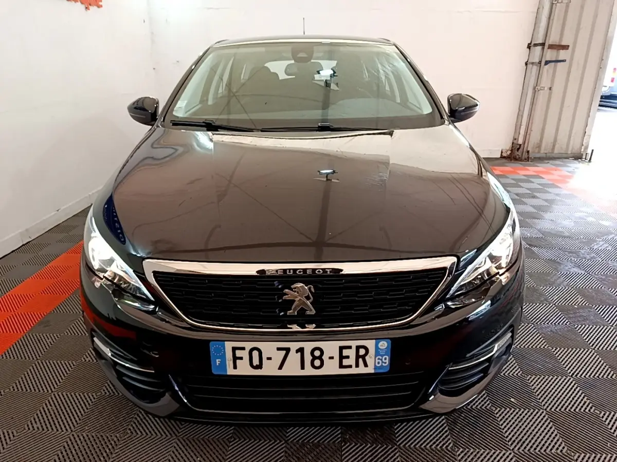 Peugeot 308 noire vue de face dans un garage, avec calandre chromée et phares allumés.