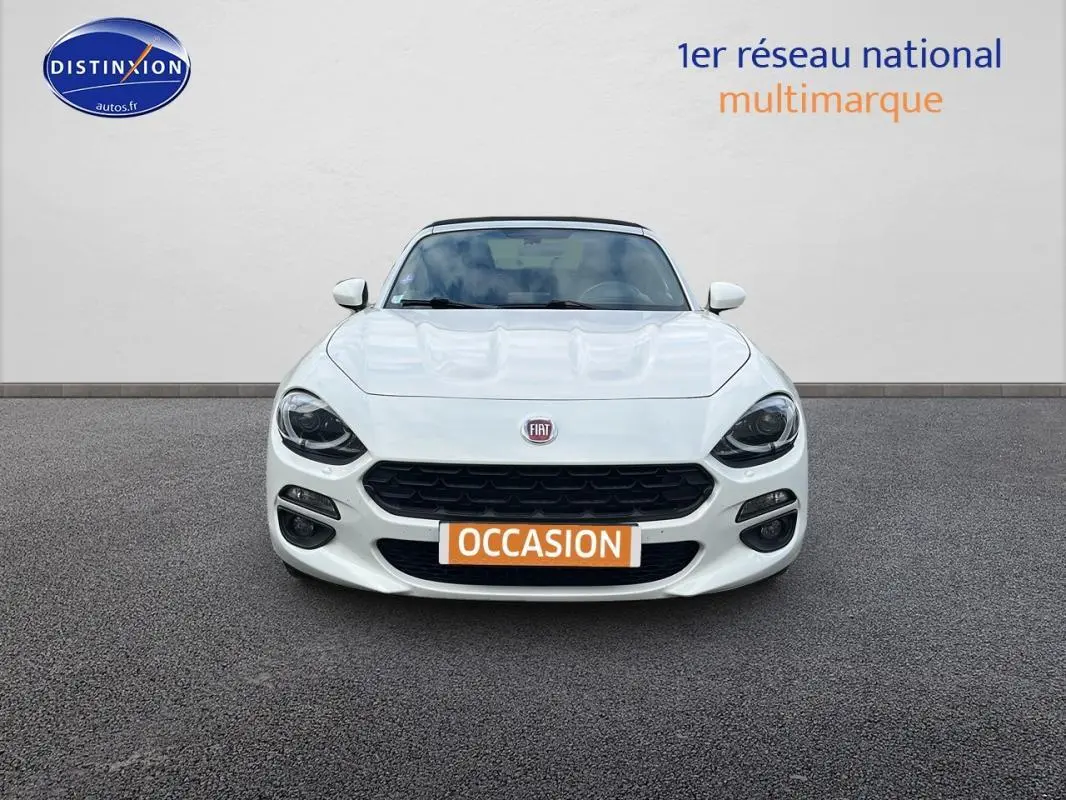 Vue de face d'une FIAT 124 Spider blanche avec phares noirs et plaque orange "OCCASION" sur un sol gris.