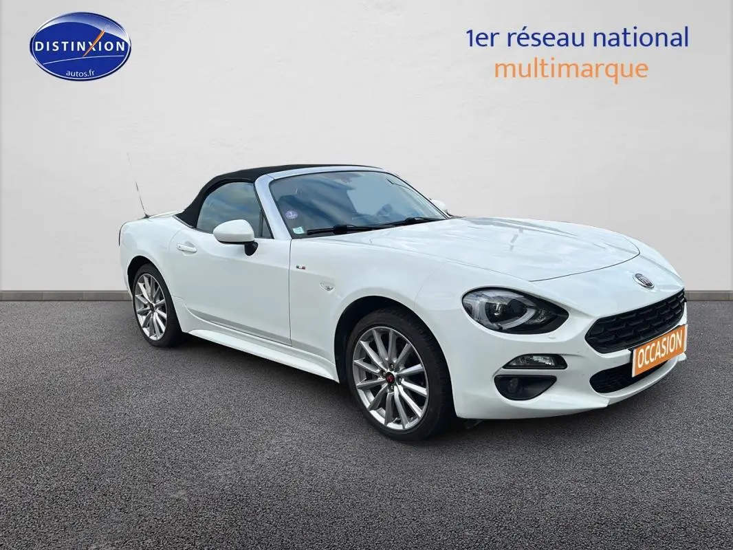Fiat 124 Spider blanc vue 3/4 avant droit, capote noire fermée et jantes alliage 17 pouces visibles.