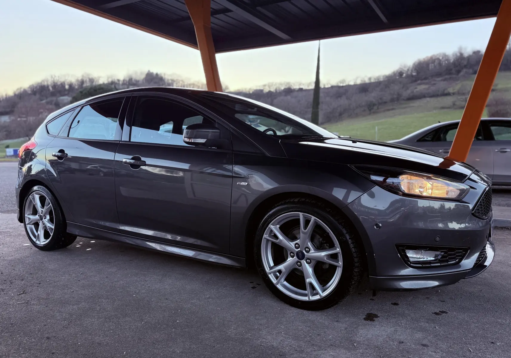 Ford Focus 1.5 TDCI ST Line grise vue de profil côté gauche sous abri, avec jantes alliage 18 pouces et phares allumés.
