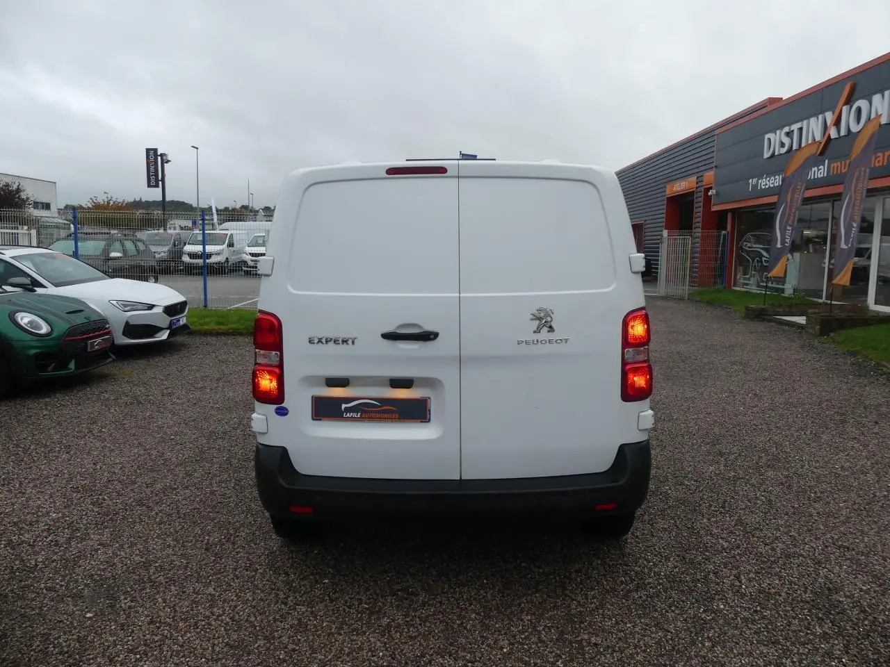 Vue arrière d'un Peugeot Expert Fourgon blanc avec feux allumés sur parking devant un bâtiment commercial.