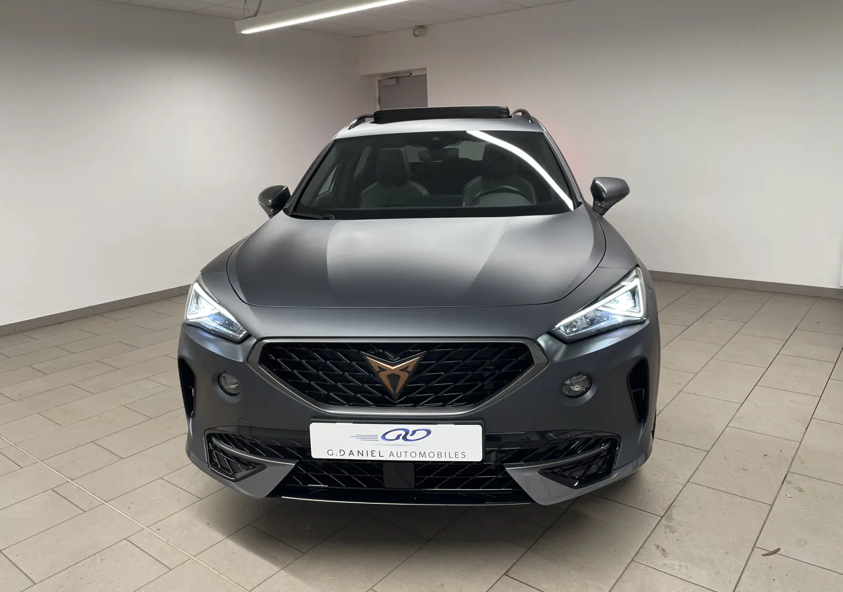 Vue avant d'un CUPRA Formentor 1.5 TSI 150 DSG7 VZ-LINE gris magnétique mat avec phares allumés en intérieur.