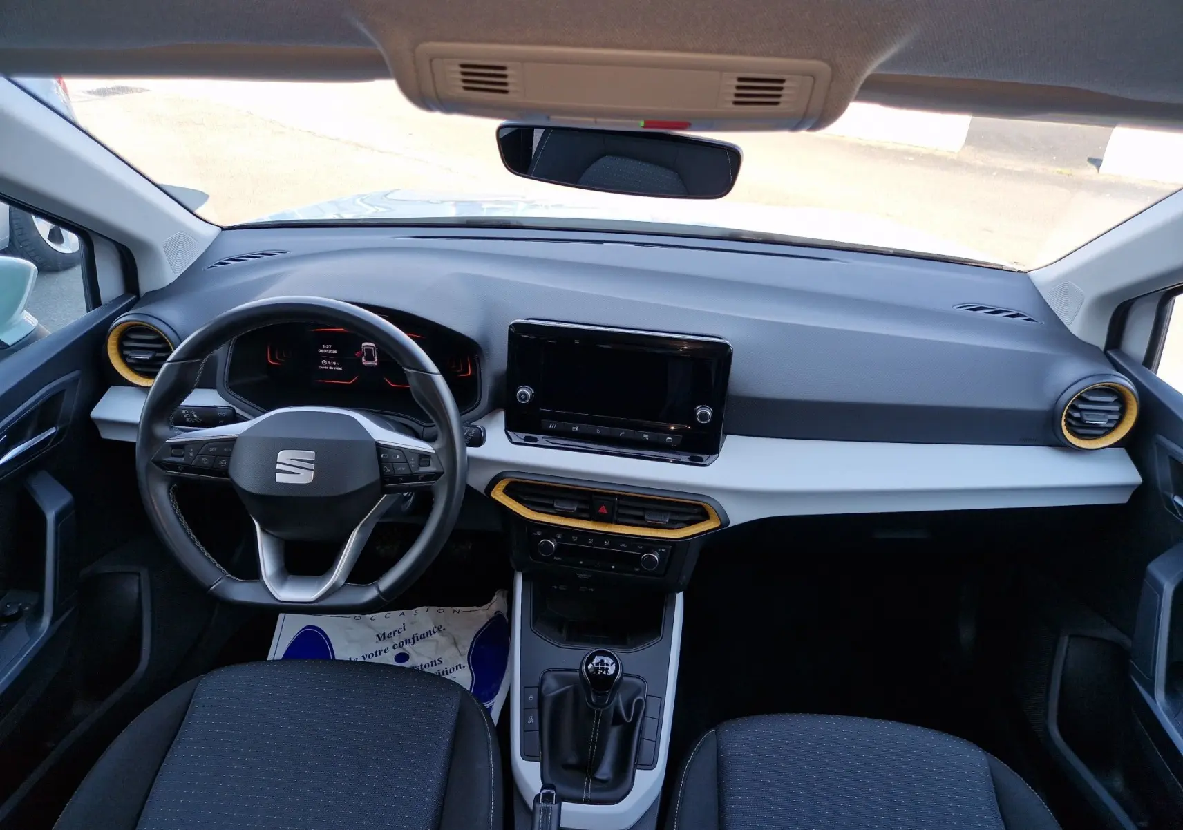 Vue intérieure avant du tableau de bord du SEAT Arona 2025, volant multifonction, console centrale avec boîte manuelle, aérateurs cerclés jaune.
