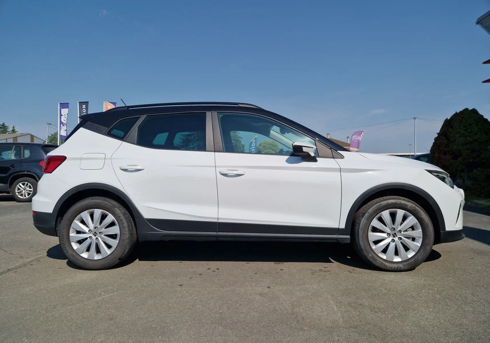 Vue de profil côté gauche d'un SEAT Arona blanc alpin avec toit noir, roues alliage et protections noires basses.