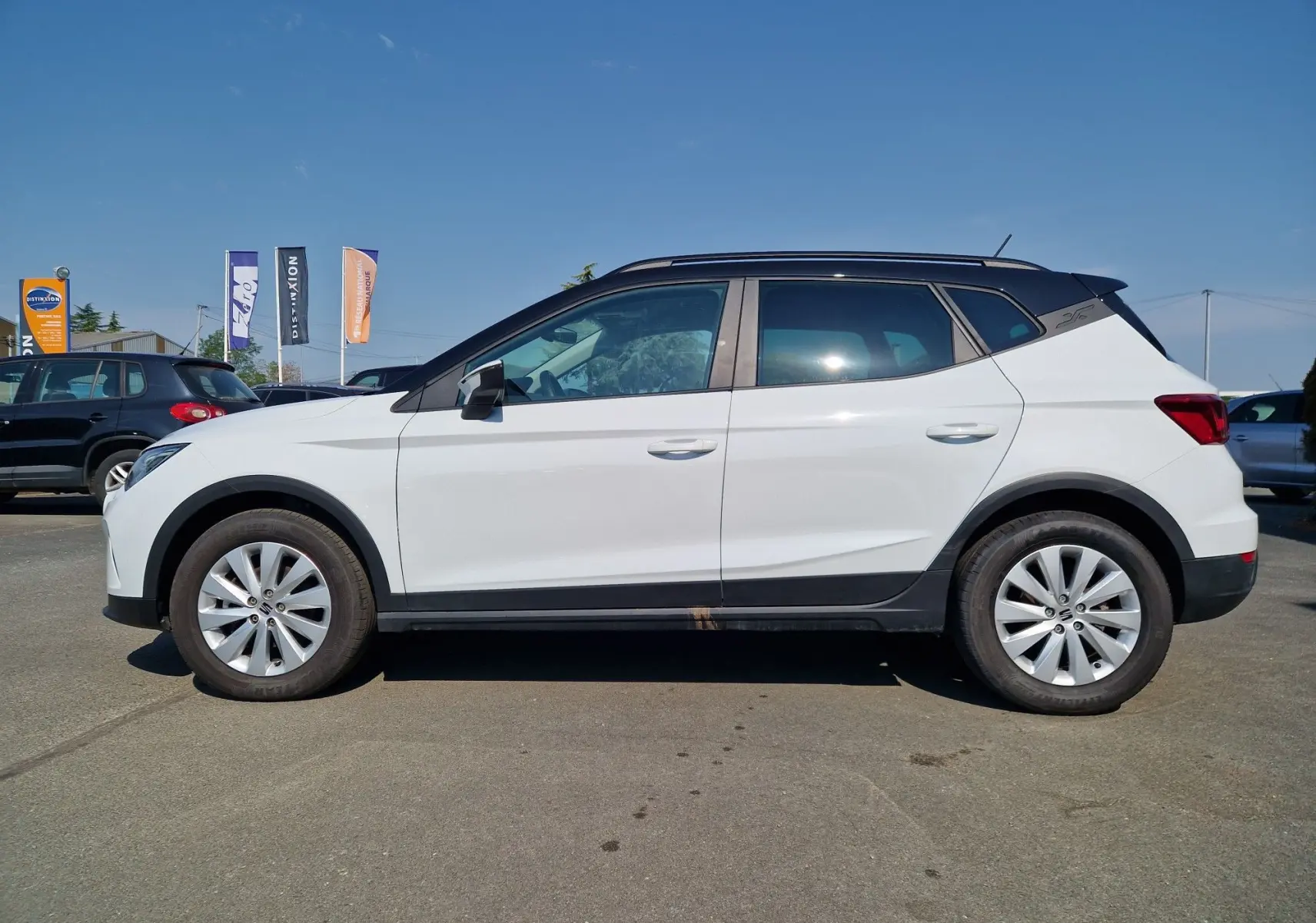 Vue de profil côté gauche d'un SEAT Arona blanc alpin avec toit noir, sur un parking en extérieur.