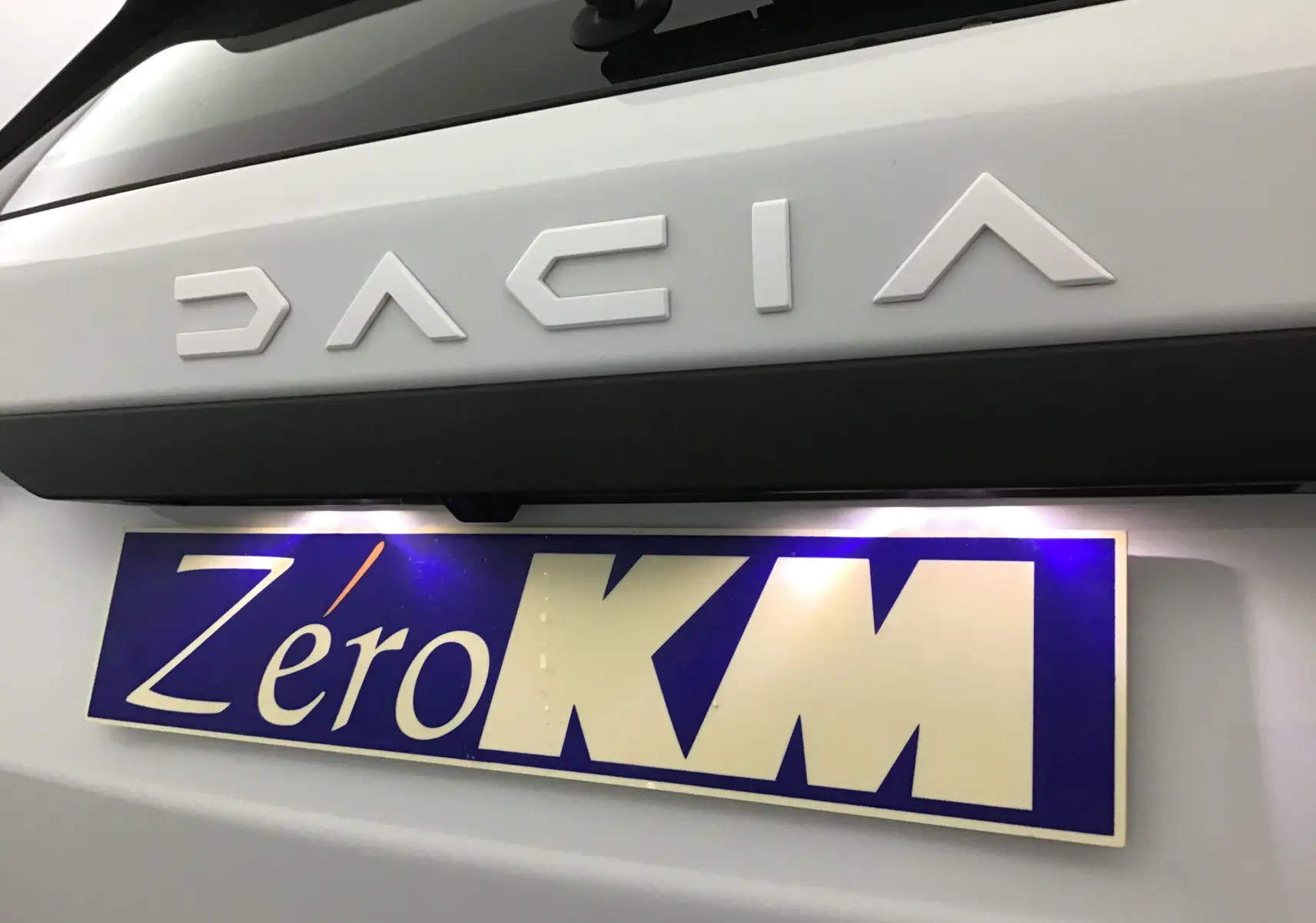 Gros plan sur le logo DACIA en relief sur la porte arrière blanche glacier du Duster 2025 avec plaque Zéro KM éclairée.