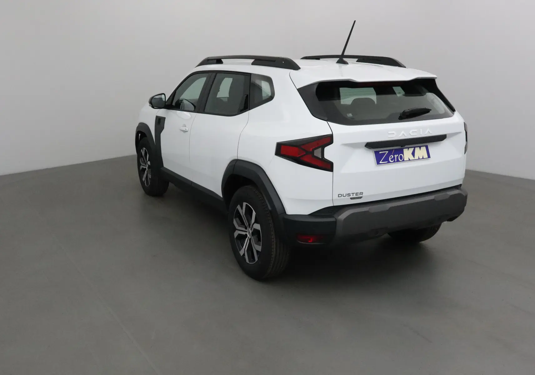Vue 3/4 arrière droite du Dacia Duster blanc glacier 2025 avec jantes alliage et barres de toit noires.