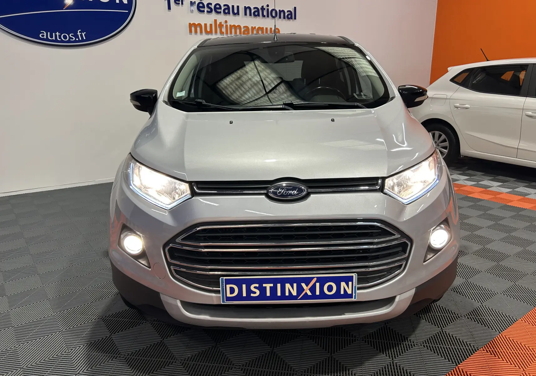 Vue avant d'un Ford EcoSport gris 2018 avec phares allumés et plaque Distinxion en intérieur showroom.