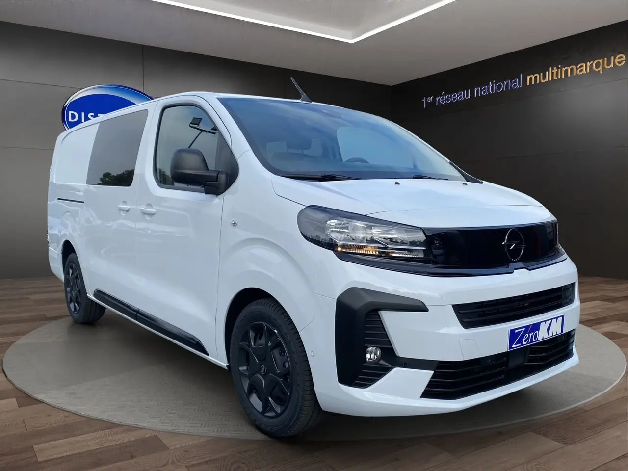 Opel Vivaro blanc vu en 3/4 avant droit, avec calandre noire et jantes foncées dans un showroom.