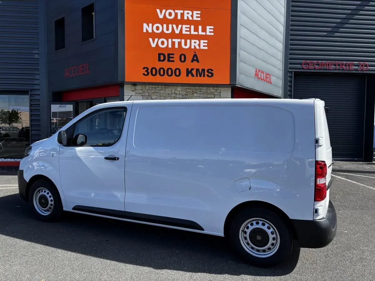 Peugeot Expert Fourgon blanc vu de profil côté gauche, stationné devant un bâtiment commercial.