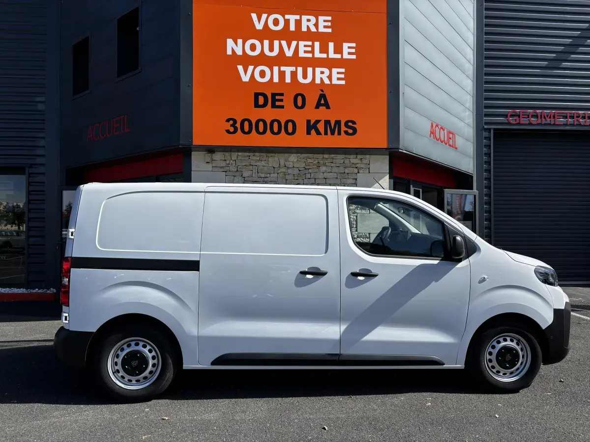 Profil droit d'un Peugeot Expert Fourgon blanc 2025 garé devant un bâtiment avec panneau orange "Votre nouvelle voiture".