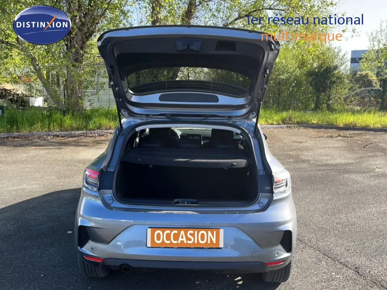 Vue arrière d'une Renault Clio gris schiste métal 2025 avec coffre ouvert, stationnée en extérieur sur sol goudronné.