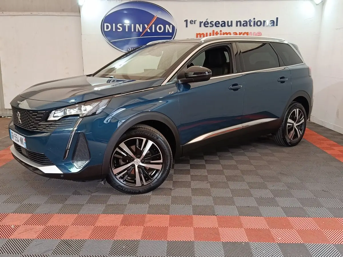 Peugeot 5008 bleu en 3/4 avant droit, avec jantes noires et détails chromés, sur sol damier intérieur showroom.
