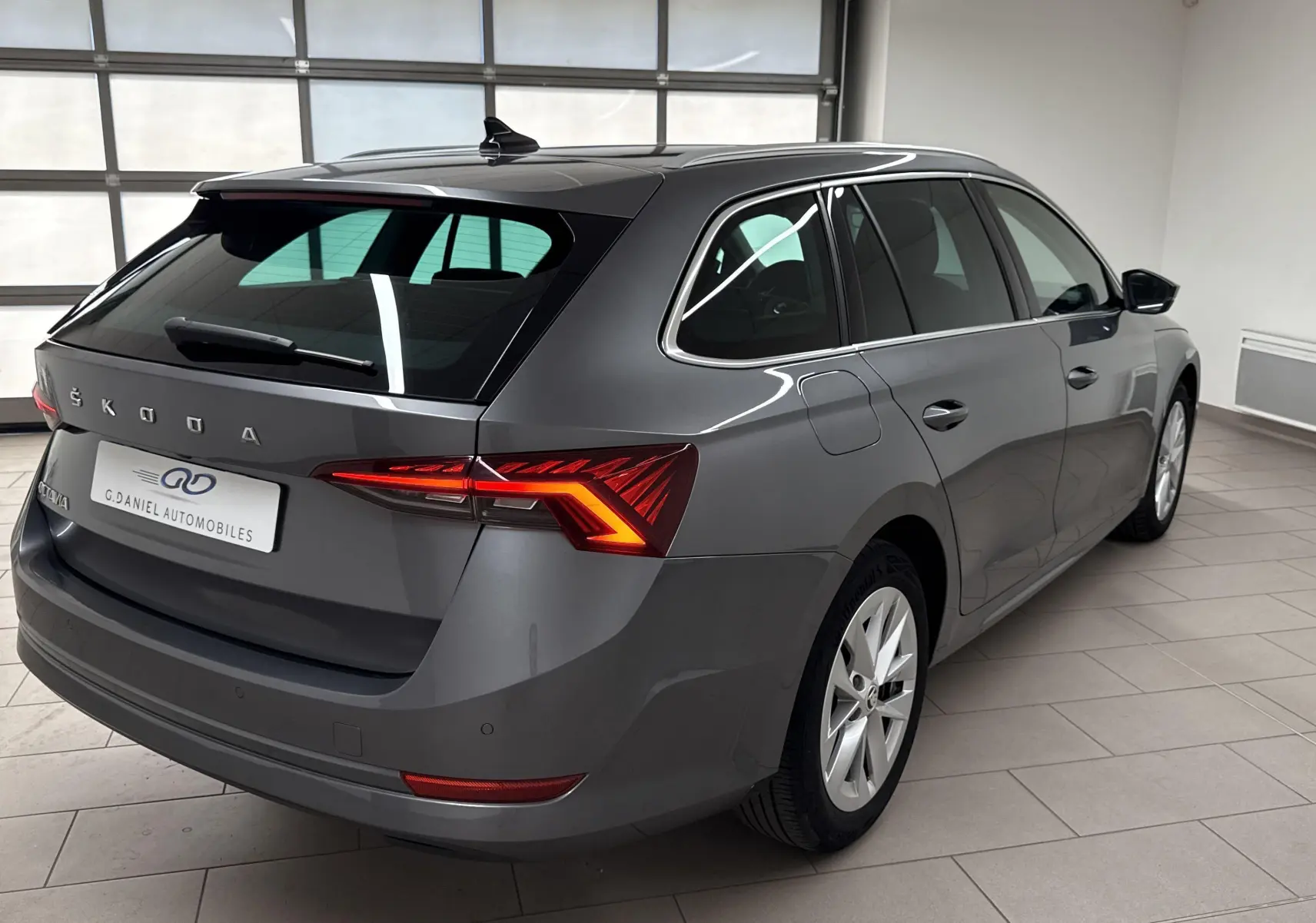 Vue 3/4 arrière droite d'une Skoda Octavia Combi 2023 gris graphite avec feux arrière allumés en intérieur showroom.