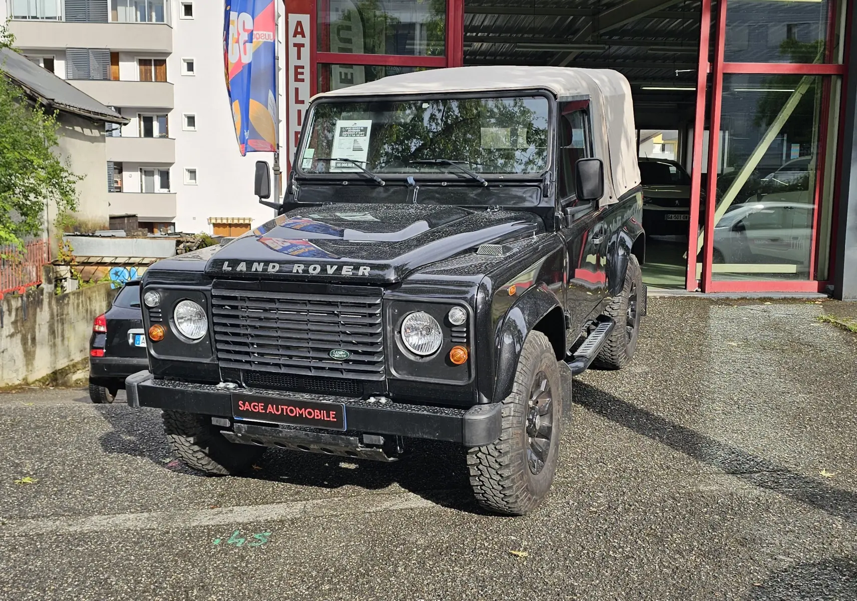Land Rover 90 Defender noir vue 3/4 avant avec toit souple beige, garé devant un garage automobile.