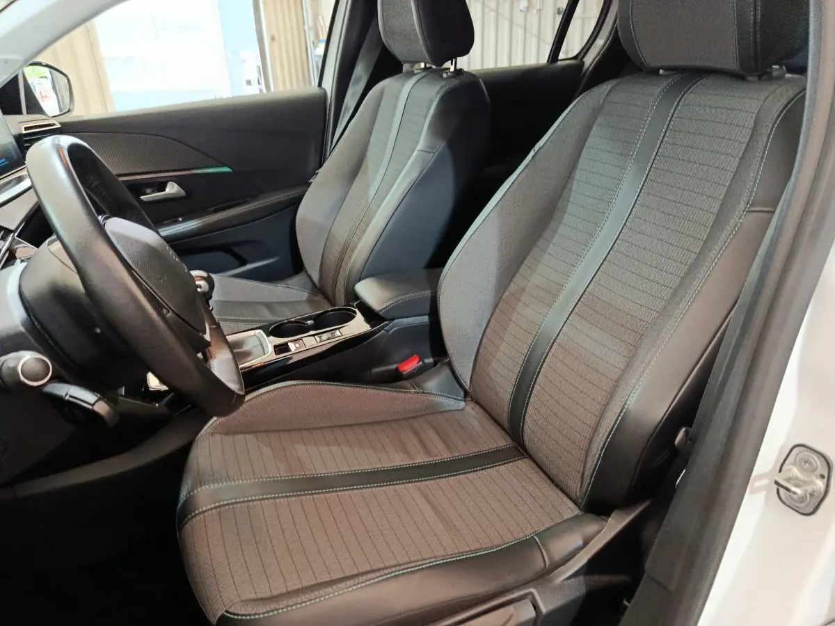 Vue intérieure côté conducteur de la Peugeot 208 blanche, mettant en valeur les sièges tissu et cuir gris avec surpiqûres vertes.