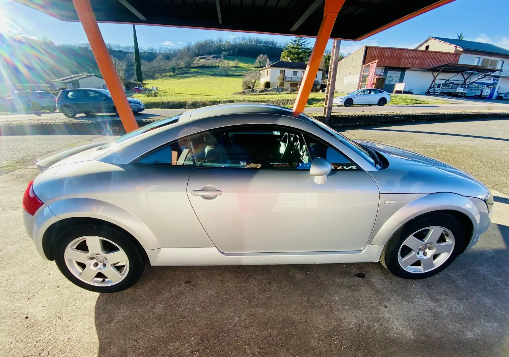 Audi TT Coupé gris vu de profil côté gauche, sous un abri avec un arrière-plan rural lumineux.