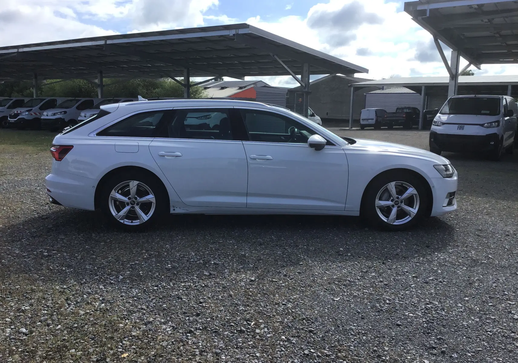 Audi A6 Avant blanc glacier vue de profil côté gauche sur un sol gravillonné avec abris de parking en arrière-plan.